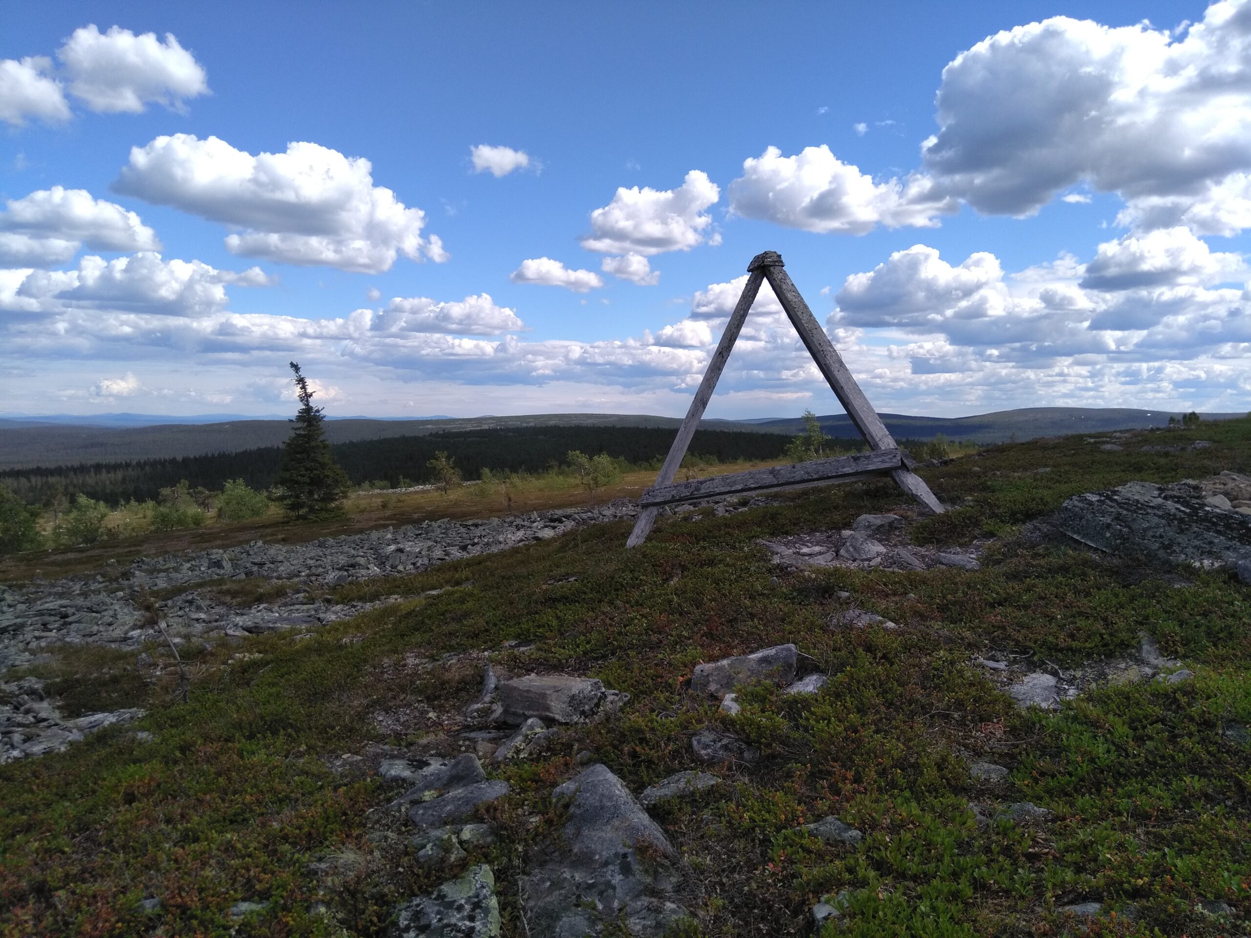 Sinisellä taivaalla on valkoisia pilviä, etualalla näkyy tunturiluontoa, jossa on puinen pyramidin mallinen kehikko